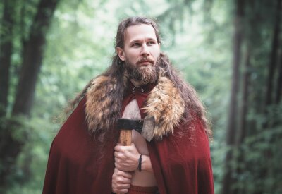 En viking med langt hår, skjegg og kappe står og ser utover skogen mens han holder en øks.