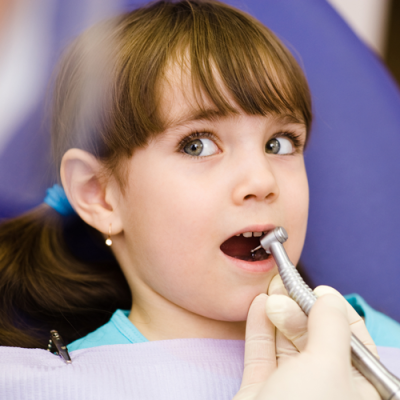 En jente som sitter og gaper i tannlegestolen, mens tannlegen holder et borr mot tennene.