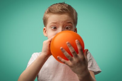 En gutt gjør store øyne mens han blåser opp en orange ballong.