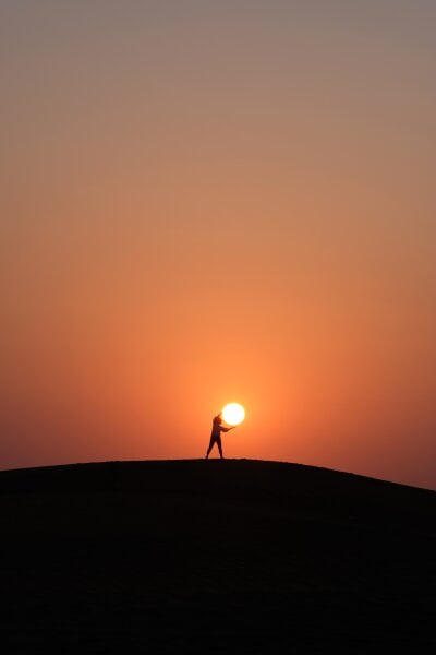 Siluett av et menneske står på toppen av an bakke med hendende utstrakt slik at det ser ut som hen "fanger" solen i solnedgangen 