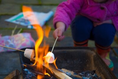 To marshmallow på pinne inn i flammene av et bål. Den ene pinnen holdes av et barn som sitter på huk